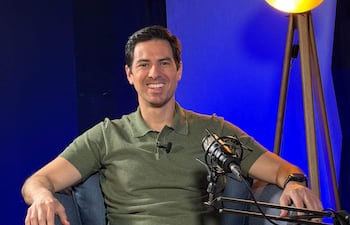 Alvaro Grau sentado en un sofá, sonriente, con camiseta verde y pantalones beige, en un ambiente de entrevista con micrófono.