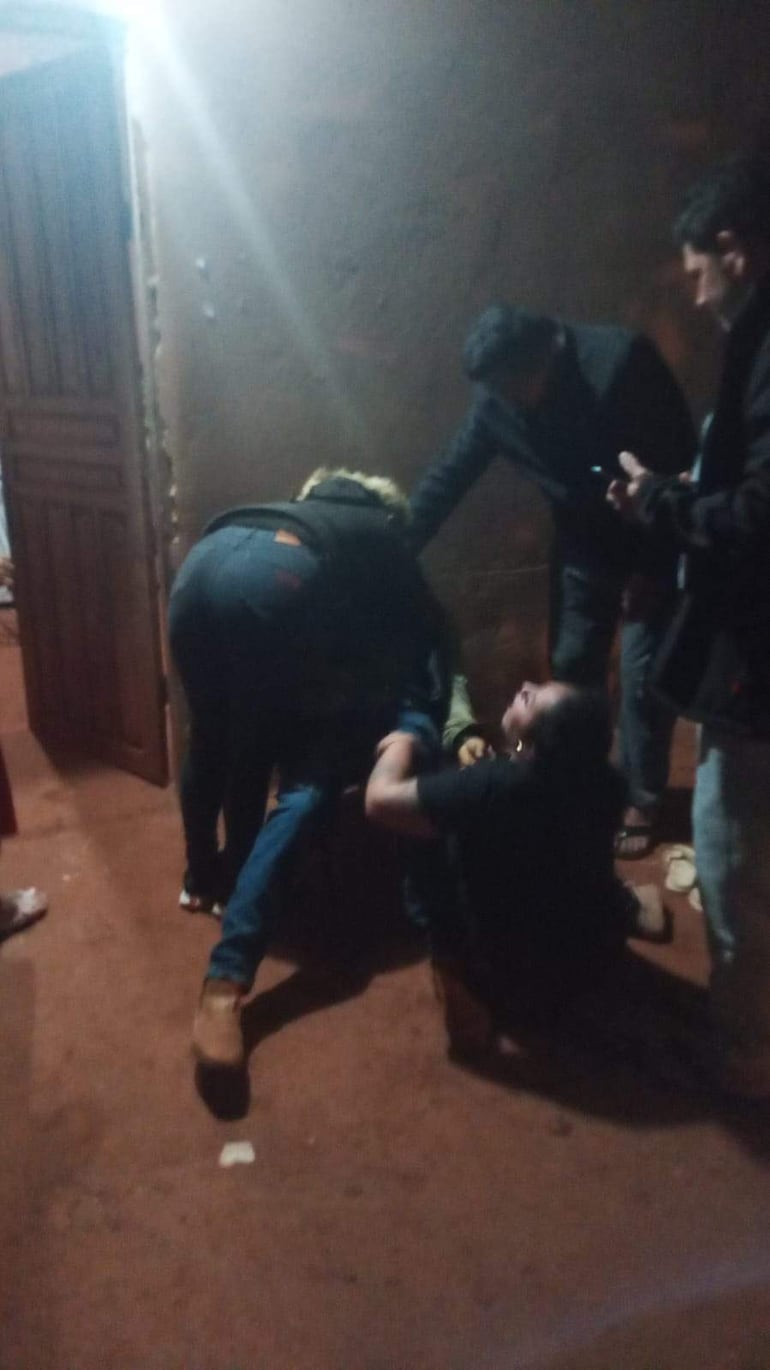 Cinco personas en una habitación con paredes marrón claro; una arrodillada, tres de pie, una con teléfono en mano, expresión de alarma.