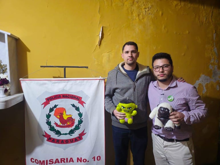 Concejales Pablo Callizo en sudadera gris y Sebastián Garay en camisa a cuadros, serios, sosteniendo muñecos de peluche en comisaría amarilla.