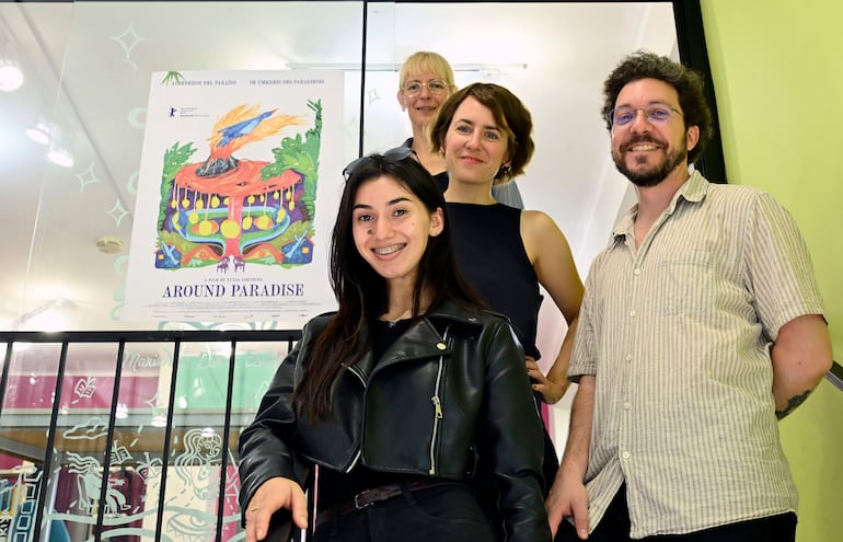 Joven sonriente con chaqueta de cuero negro, dos mujeres y un hombre en un evento cultural con cartel de película colorido detrás.