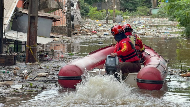 Dos rescatistas con chalecos y cascos navegan en un bote rojo en un área inundada y llena de desechos.