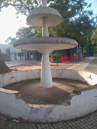 Fuente de tres niveles en mal estado, rodeada de árboles y sillas rosas en un espacio vacío, sin personas presentes.