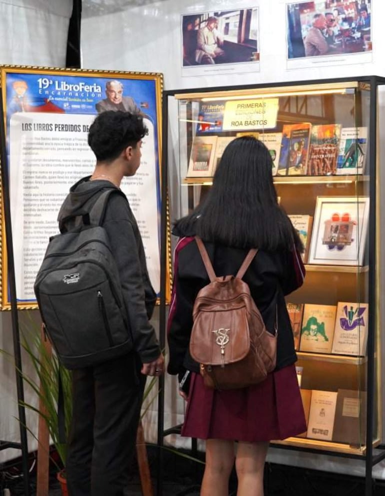 Mujer de abrigo oscuro y hombre con camiseta oscura y mochila observan libros en una vitrina en un evento cultural.