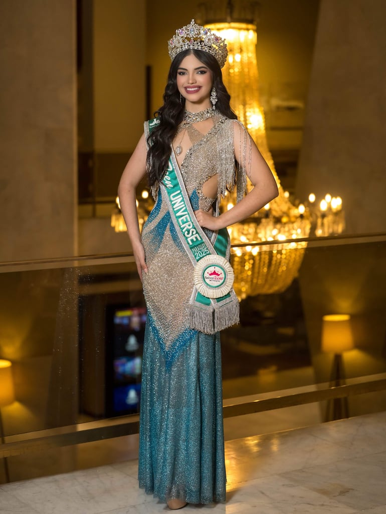 KatyaJi de pie, con corona y vestido brillante azul y plateado, en un evento elegante con lámpara de cristal detrás.