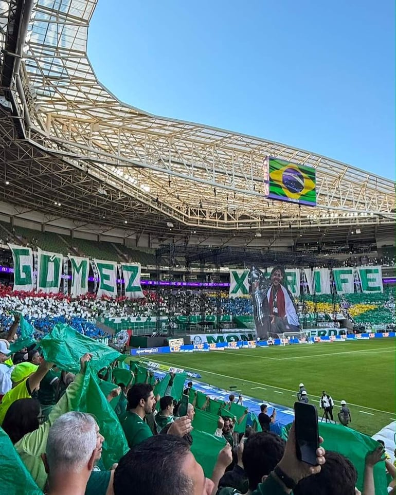 Aficionados con banderas verdes en estadio, ambiente festivo con pancartas de apoyo y pantalla mostrando trofeo.