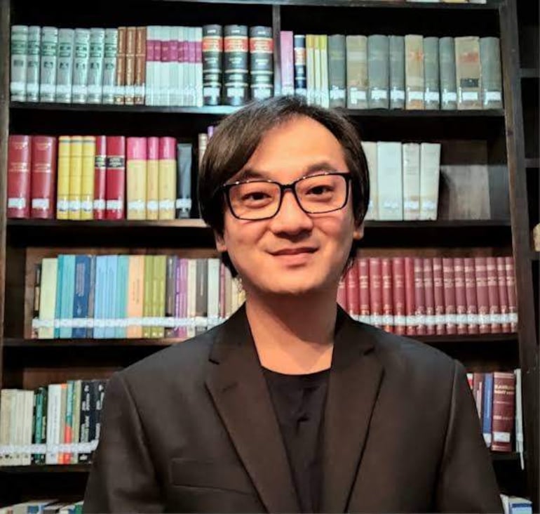 Deny Yoon Pak con gafas y atuendo negro frente a una estantería repleta de libros, sonriendo levemente.