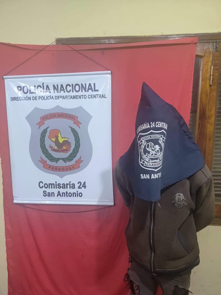 Hombre cubierto con paño azul oscuro en comisaría, junto a cartel de Policía Nacional y fondo rojo.
