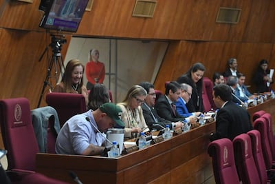 Mujer de pie sonriendo en el fondo, varias personas en vestimenta formal, revisando teléfonos y conversando en un ambiente institucional.