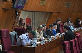 Mujer de pie sonriendo en el fondo, varias personas en vestimenta formal, revisando teléfonos y conversando en un ambiente institucional.