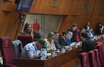 Mujer de pie sonriendo en el fondo, varias personas en vestimenta formal, revisando teléfonos y conversando en un ambiente institucional.