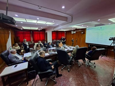 Mujer de pie presenta en sala de reuniones formal, asistentes atentos con documentos en manos y pantalla proyectando información.