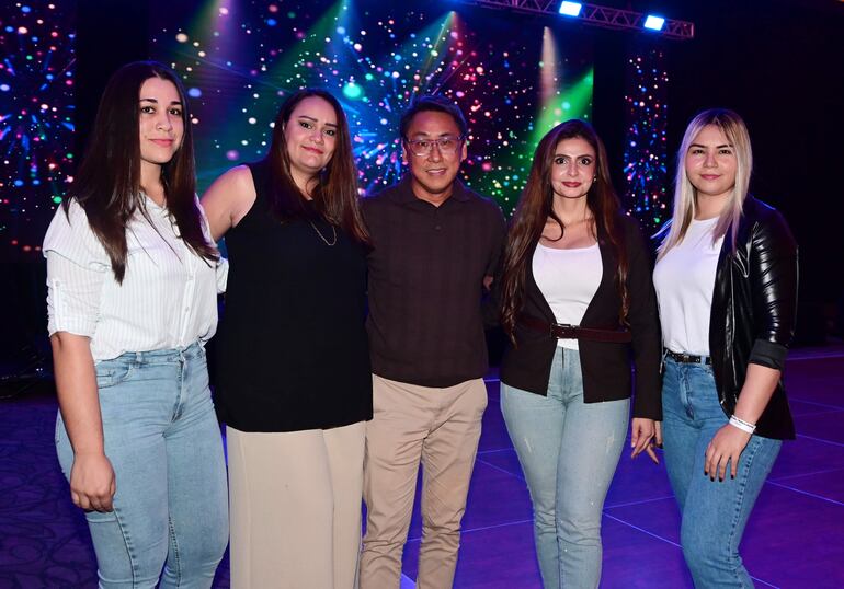 Cinco personas sonríen en ambiente festivo; mujeres con vestidos y chaquetas, hombre en camisa marrón, luces brillantes de fondo.