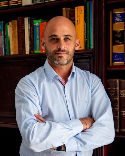 Manuel Guanes Nicoli, de pie con camiseta azul clara, frente a estantería repleta de libros, con expresión tranquila.