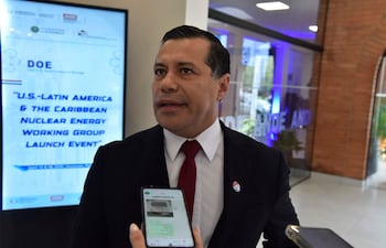 Félix Sosa con traje negro y corbata roja sostiene un teléfono, en un ambiente formal con una pantalla de fondo.