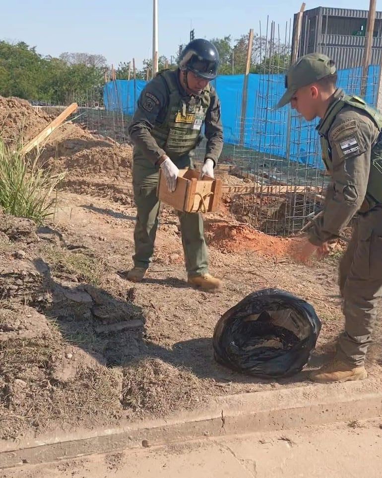 Dos trabajadores en uniformes verdes y guantes, uno sosteniendo una caja mientras el otro revisa una bolsa negra en un terreno desenterrado.