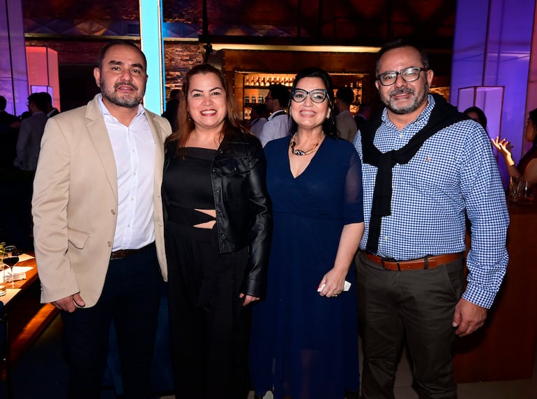 Hombres y mujeres sonrientes en un evento, con chaquetas y vestidos, en un ambiente moderno con bar al fondo.