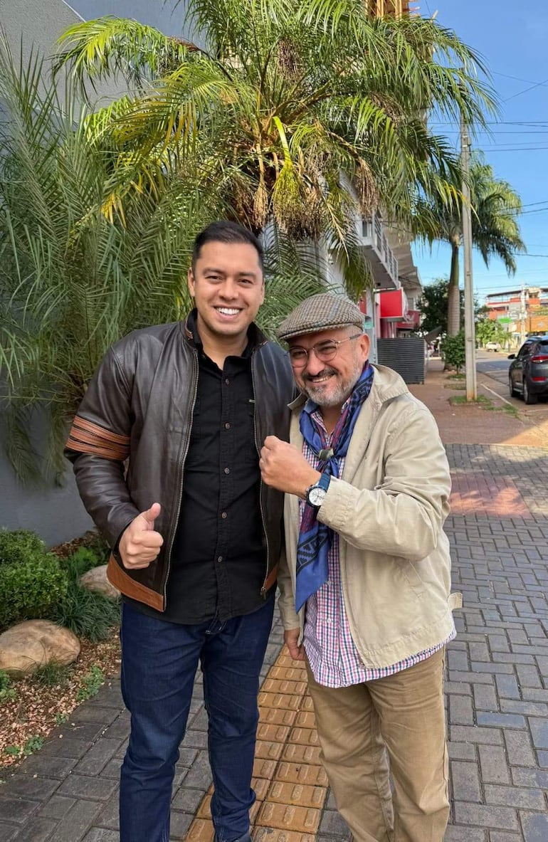 Luis Yd con chaqueta de cuero y Miguel Prieto Vallejos en abrigo claro, ambos sonriendo en una calle con palmeras al fondo.