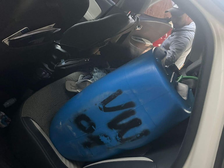 Hombre con gorra y suéter gris en un vehículo, rodeado de un barril azul y objetos dispersos.