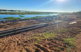 Terreno con tuberías y materiales de construcción en un paisaje natural junto al río, bajo un sol brillante.