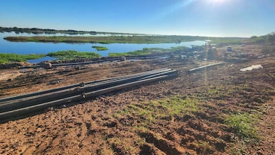 Terreno con tuberías y materiales de construcción en un paisaje natural junto al río, bajo un sol brillante.
