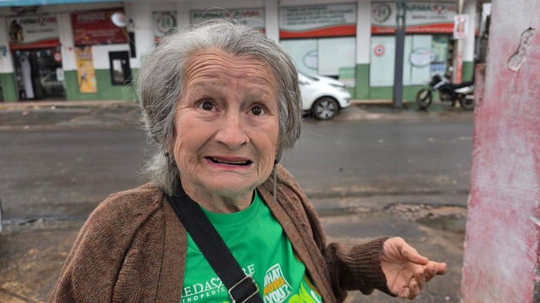 María Gutiérrez, mujer mayor con cabello canoso y camiseta verde, gesticula con preocupación en un entorno urbano lluvioso.