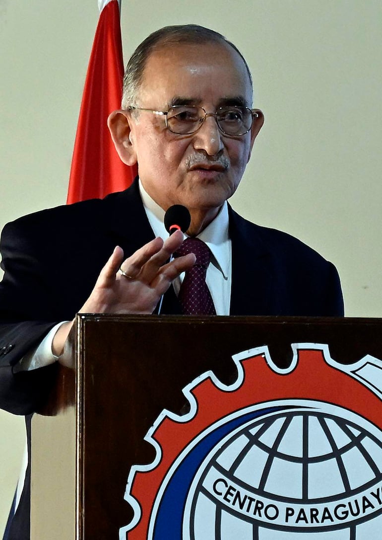 Orador de cabello canoso y gafas, con mano levantada, detrás de podio con logo del 'Centro Paraguay' en rojo y negro.