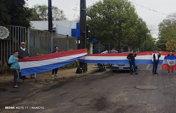Grupo de seis personas en protesta, sosteniendo banderas rojas, blancas y azules, con agentes de seguridad al fondo.