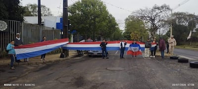 Grupo de seis personas en protesta, sosteniendo banderas rojas, blancas y azules, con agentes de seguridad al fondo.