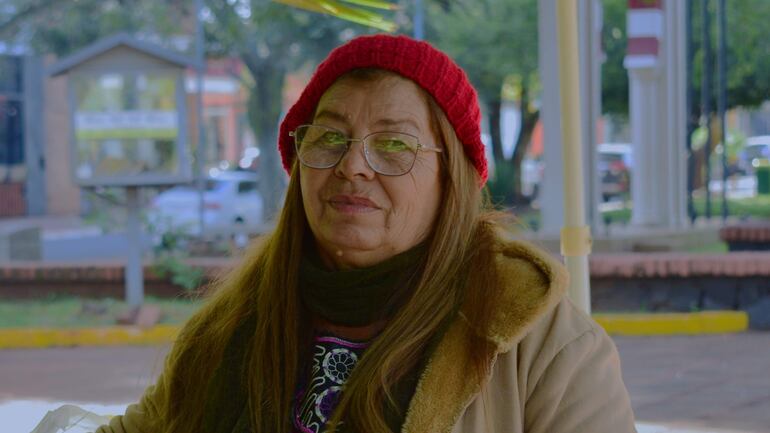 Evelia Silva sentada con gorro rojo y gafas, cabello largo suelto, chaqueta clara y bufanda oscura, en ambiente urbano.