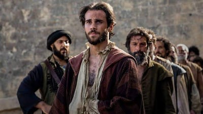 Hombre con cabello oscuro y barba, vestido con ropas de época, rodeado por otros hombres en un ambiente rústico.