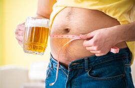 Un hombre se mide la circunferencia de la panza con una mano, mientras con la otra sostiene un chopp de cerveza.