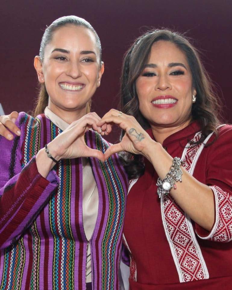 Dos mujeres sonrientes, una con atuendo colorido y trenzas, otra en rojo, hacen un gesto de corazón con las manos en un ambiente oscuro.