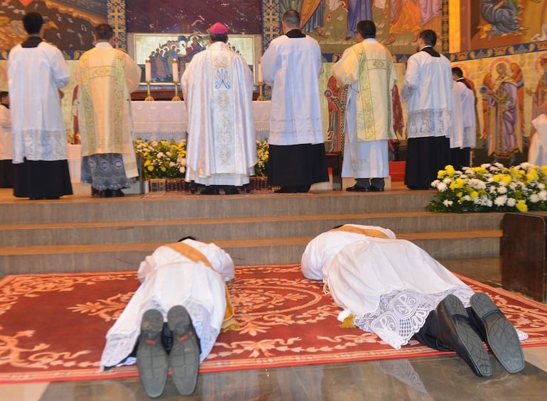 Dos hombres realizan acto de postración en el altar, rodeados de sacerdotes con vestiduras litúrgicas y un ambiente decorado.