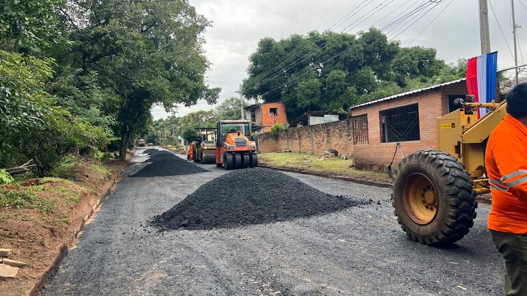 Trabajador con chaleco naranja supervisa maquinaria pesada en la reparación de una calle rodeada de casas y árboles.