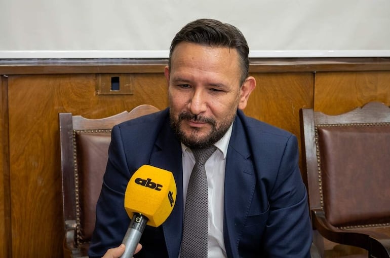 Derlis Léon con barba y traje oscuro, sosteniendo un micrófono amarillo en un espacio con paredes de madera.