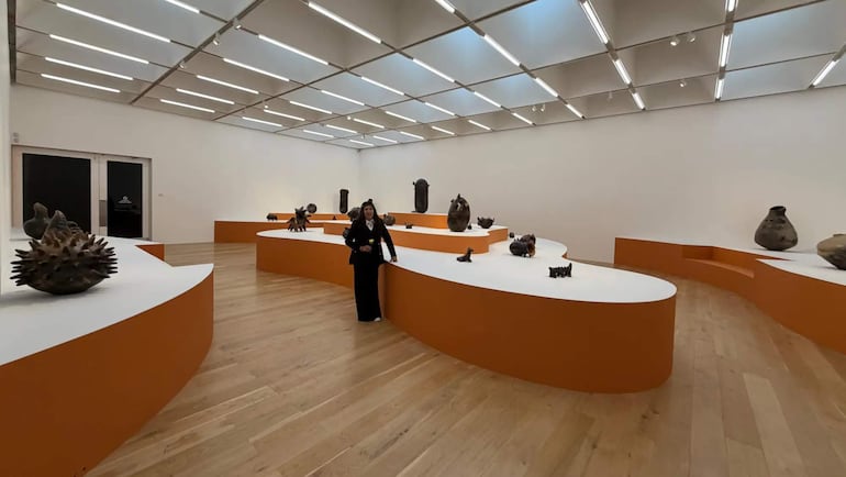 Julia Isídrez, con vestido oscuro y cabello recogido, observa esculturas en un museo con iluminación brillante y mesas curvadas naranjas.