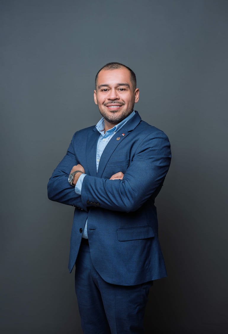 Hombre en traje azul oscuro sonriente, brazos cruzados sobre el pecho, con fondo gris y postura confiada.