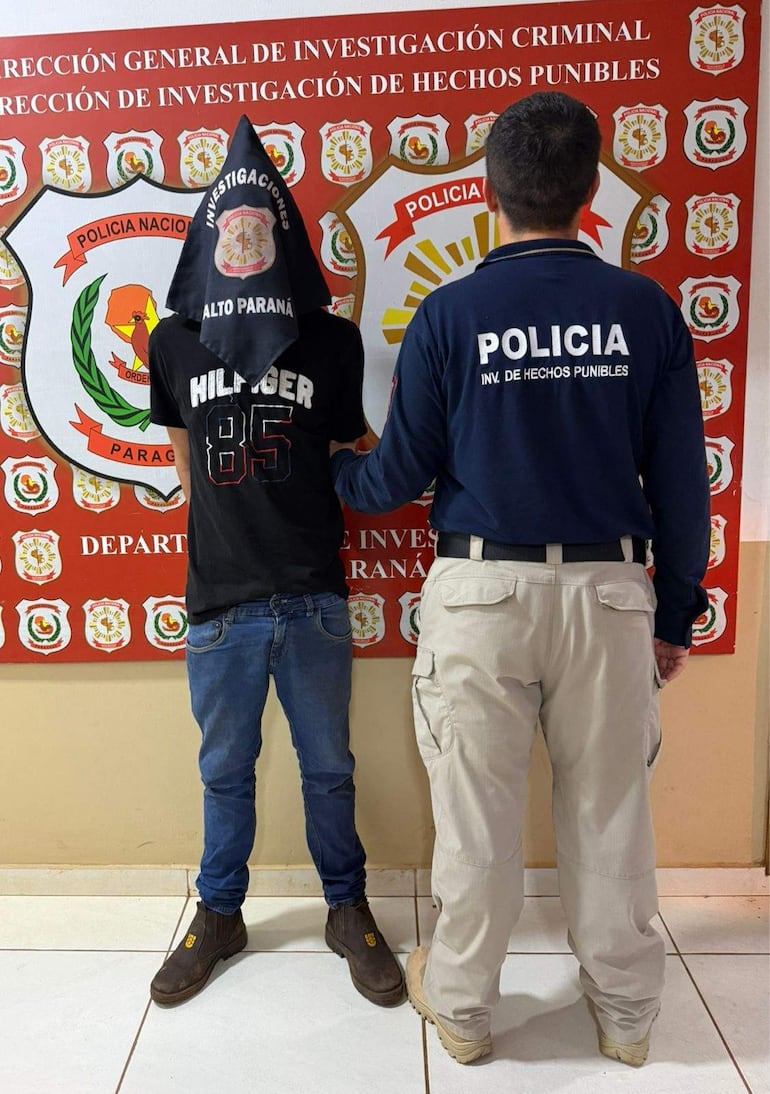 Hombre con camisa negra y pañuelo en el rostro, junto a un policía en uniforme oscuro en una comisaría.