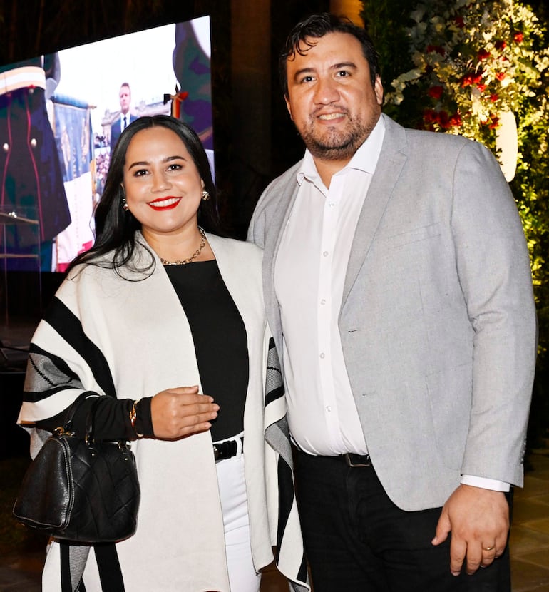 Mujer con blusa negra y poncho blanco sostiene un bolso negro, junto a un hombre de chaqueta gris y camisa blanca, sonriendo en evento.