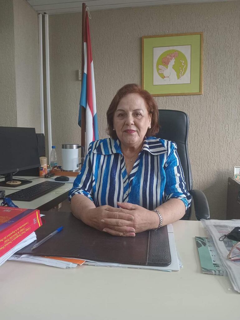 Lici Teresita Sánchez, con blusa a rayas, revisa documentos en su escritorio, rodeada de botellas de agua y una computadora.