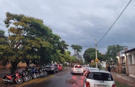 Calle urbana con coches y motos estacionados bajo un cielo nublado y lluvia, pavimento mojado refleja el entorno.