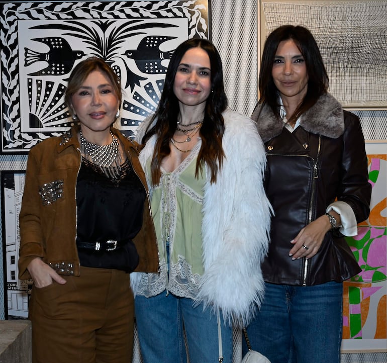 Tres mujeres posan en un entorno artístico. Dos llevan chaquetas, una viste de piel, sonriendo en un ambiente moderno.