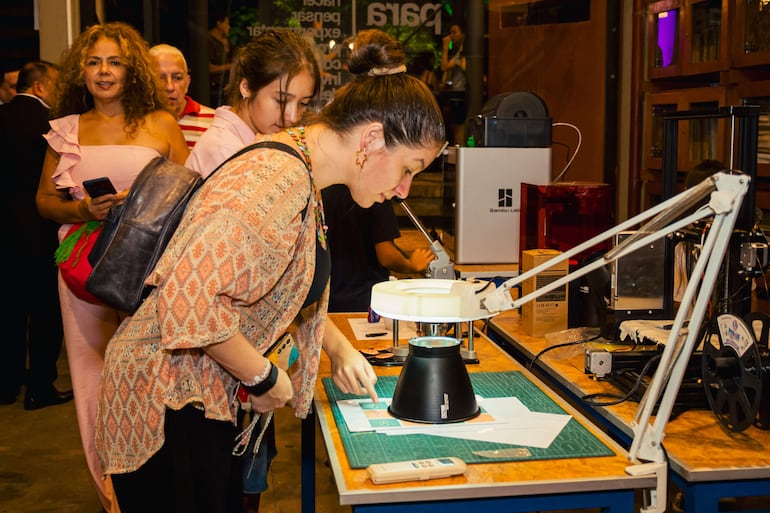 Mujer de cabello castaño usa lupa en mesa con herramientas, otra mujer mira al frente sosteniendo un móvil, ambiente moderno y tenue.