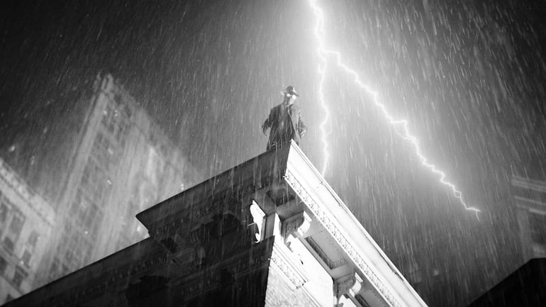 Cage con abrigo y sombrero, de pie al borde de un edificio, bajo intensos rayos y lluvia en gris.