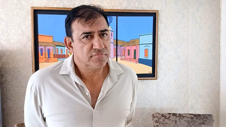 Hombre en camisa blanca con canas, de pie frente a pintura de casas coloridas, con expresión seria y postura inclinada.