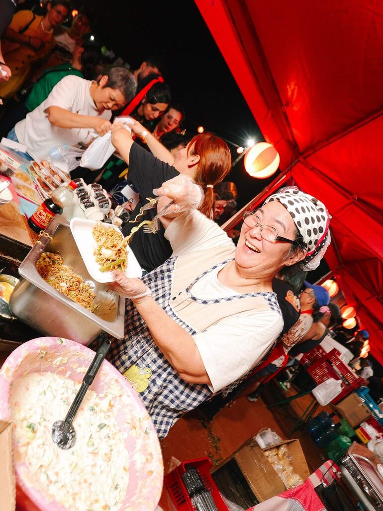 Mujer canosa con delantal a cuadros sonríe mientras sirve noodles, rodeada de un animado ambiente festivo.