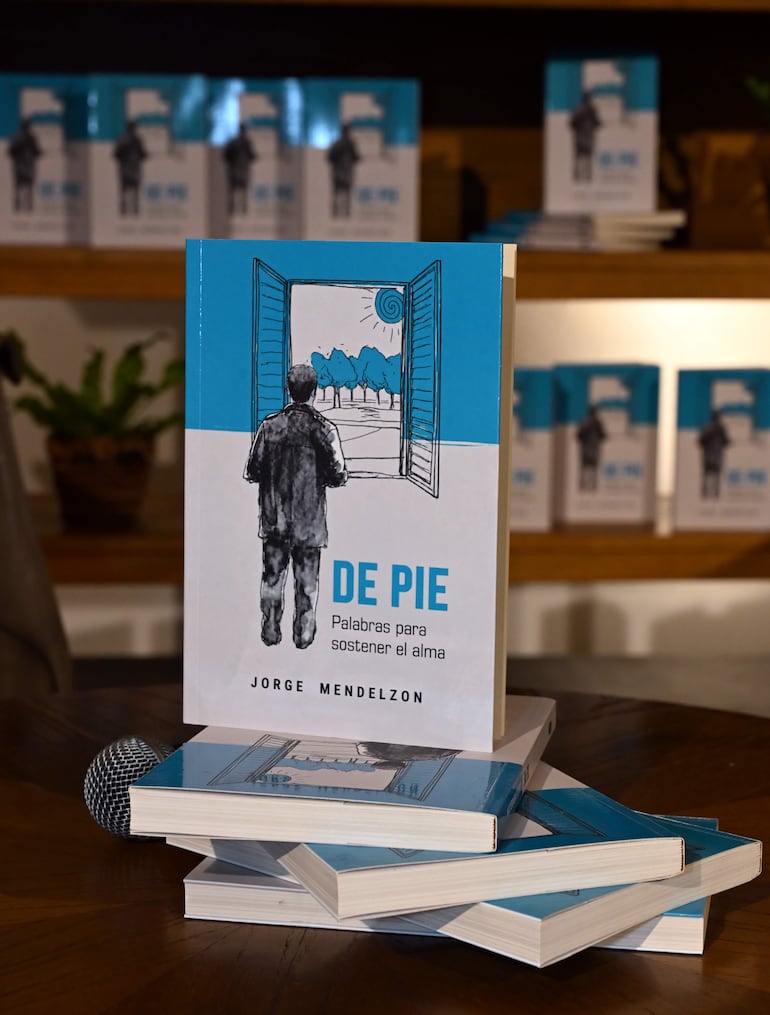 Varios ejemplares del libro 'De pie' apilados sobre una mesa de madera, con un micrófono al frente y estantería de fondo.