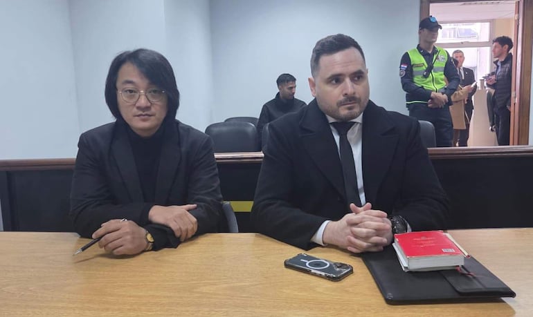 Deny Yoon Pak y Silvio Corbeta Dinamarca, en trajes oscuros, esperan en mesa con posturas atentas en sala de audiencia.