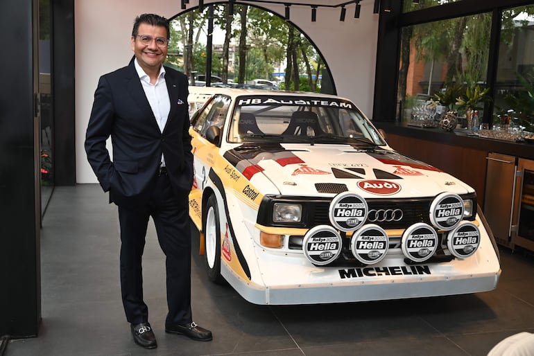 Hombre sonriente en traje oscuro posando junto a un automóvil Audi decorado con logotipos de patrocinadores en un ambiente elegante.