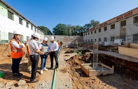 Mujer con casco naranja observa mientras hombres revisan documentos en un sitio de construcción bajo un cielo claro.